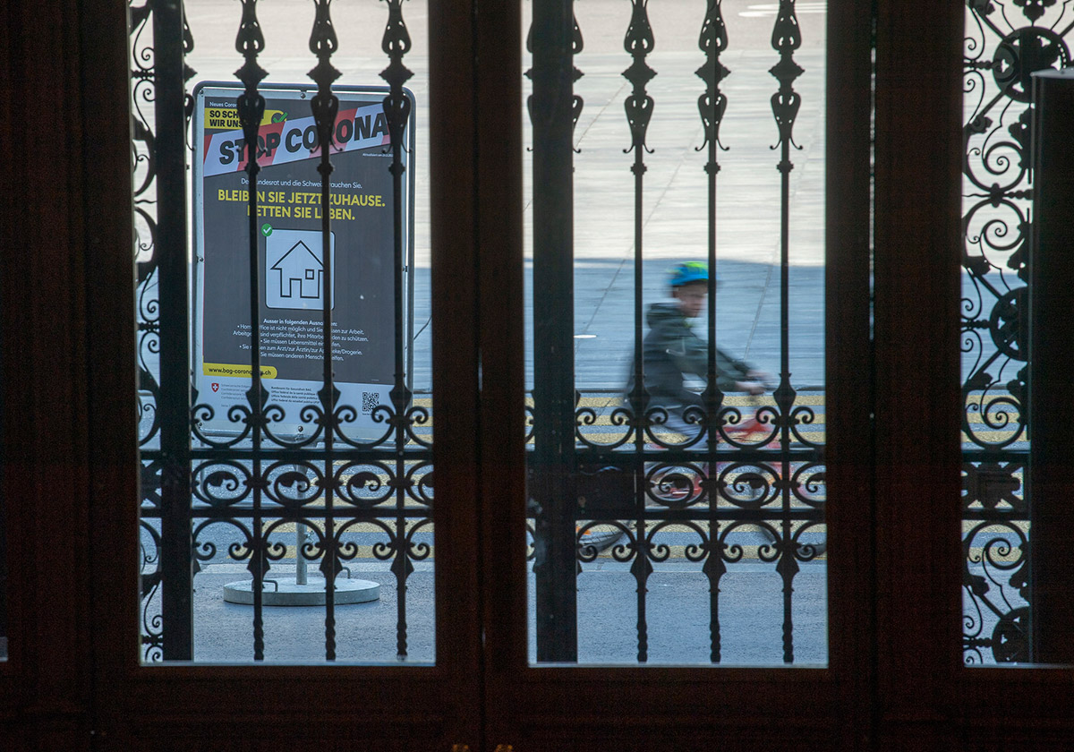 Vor dem Bundeshaus fordert ein Plakat der ‹Stop-Corona-Kampagne› die Bevölkerung dazu auf zuhause zu bleiben. Es ist durch das schmiedeeiserne Gitter vor dem Haupteingang des Bundeshauses aufgenommen.